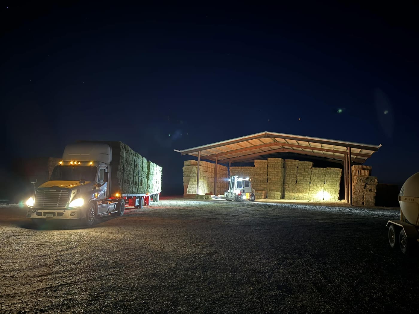 A semi loaded with hay at night, parked beside an open shed where a forklift sits among stacked bales