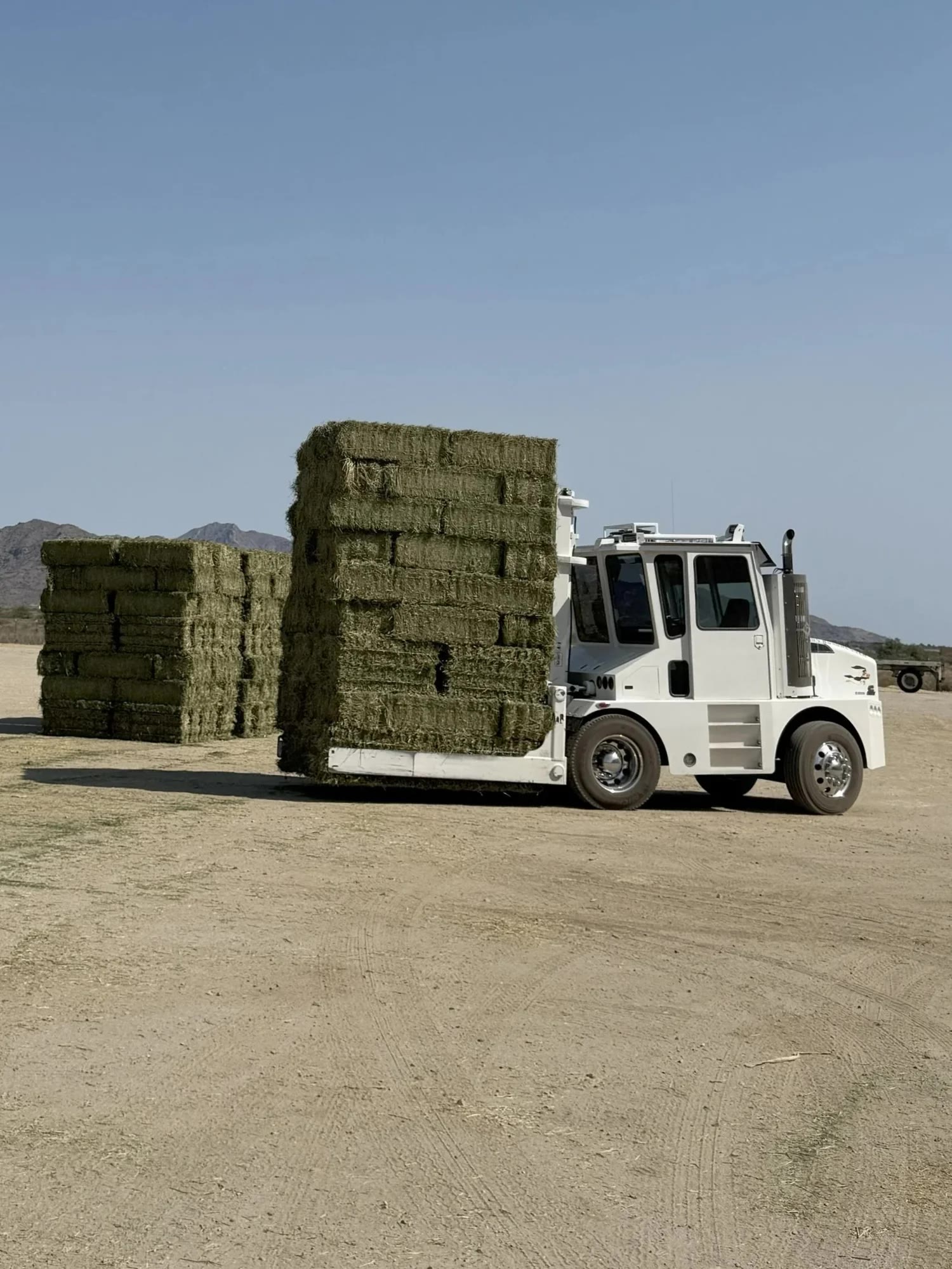 A white loader carrying a tall stack of green hay bales in a dirt yard, with another stack and desert mountains behind